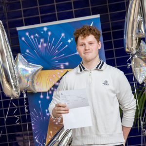 Harry Finnigan holding his T Level results stood in front of a blue banner next to some silver baloons.