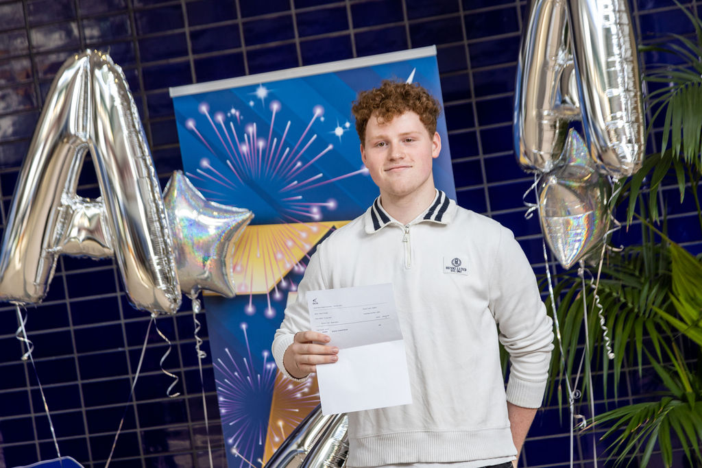 Harry Finnigan holding his T Level results stood in front of a blue banner next to some silver baloons.