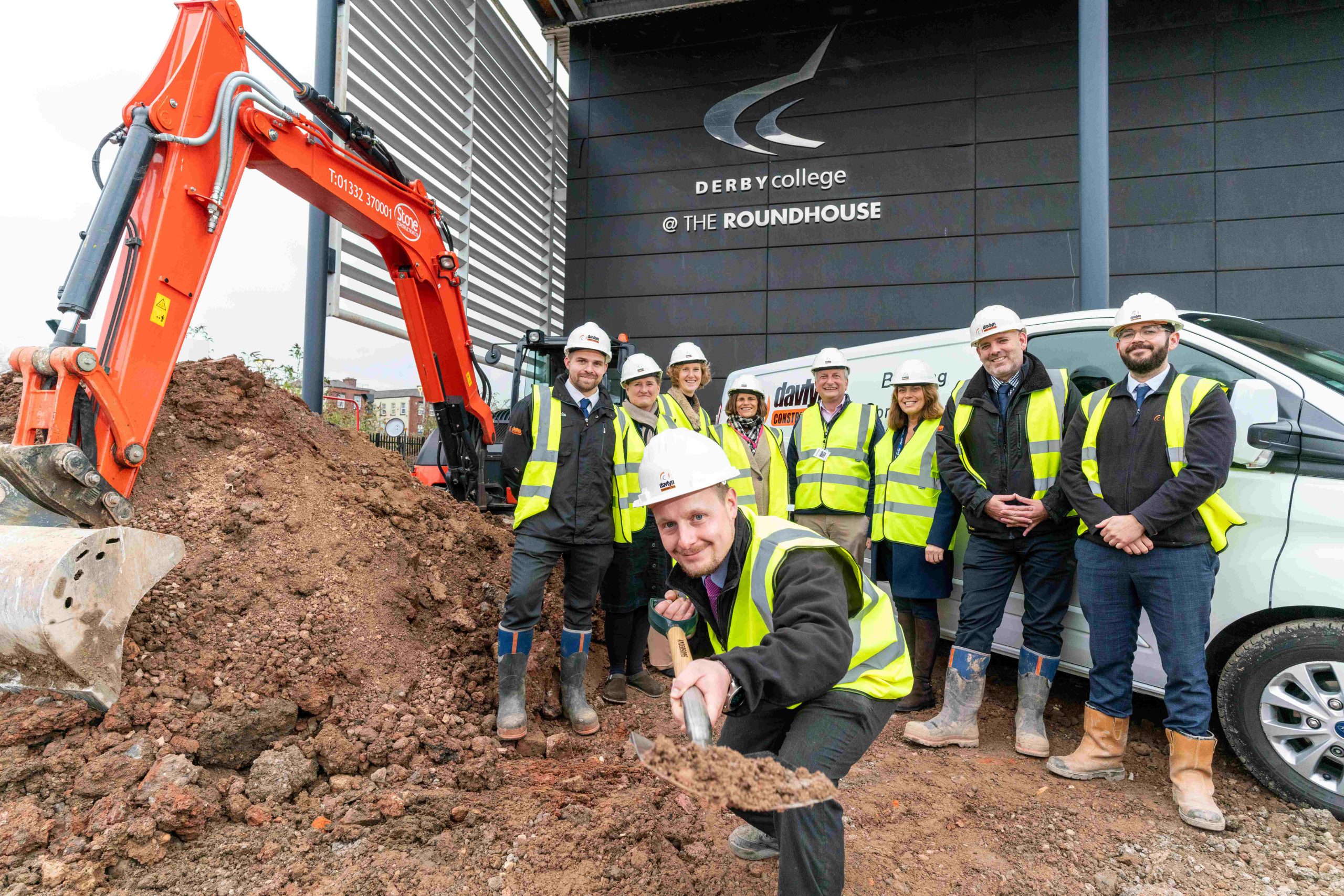 Man holding a shovel with dirt stood in front of a group of people and a digger.