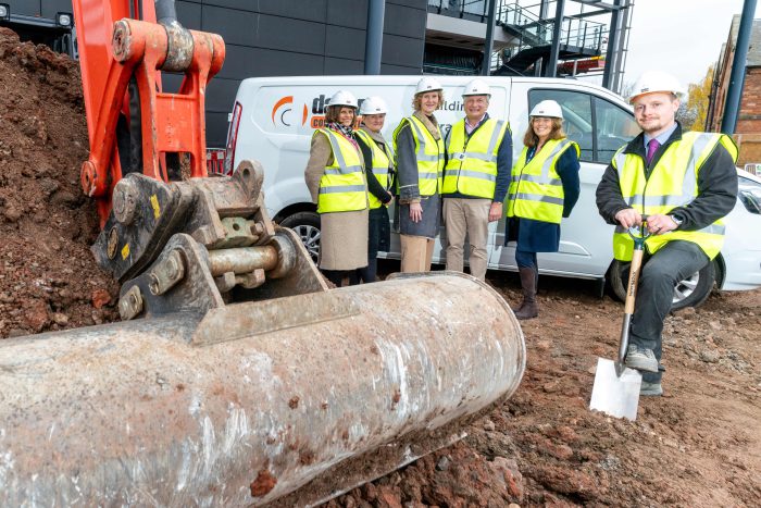 Man holding a shovel that is dug into the ground next to a digger.