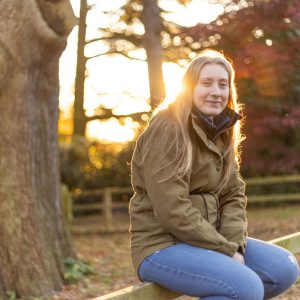 Young lady sat on a wooden fence by a tree.