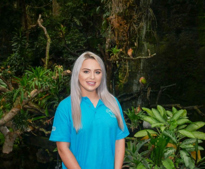 Female apprentice stood in front of trees, bushes and foliage.