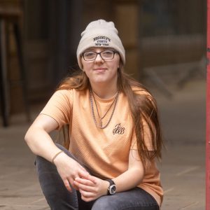 Young girl with glasses wearing a beanie hat and orange t-shirt sat with her arms rested on her legs.