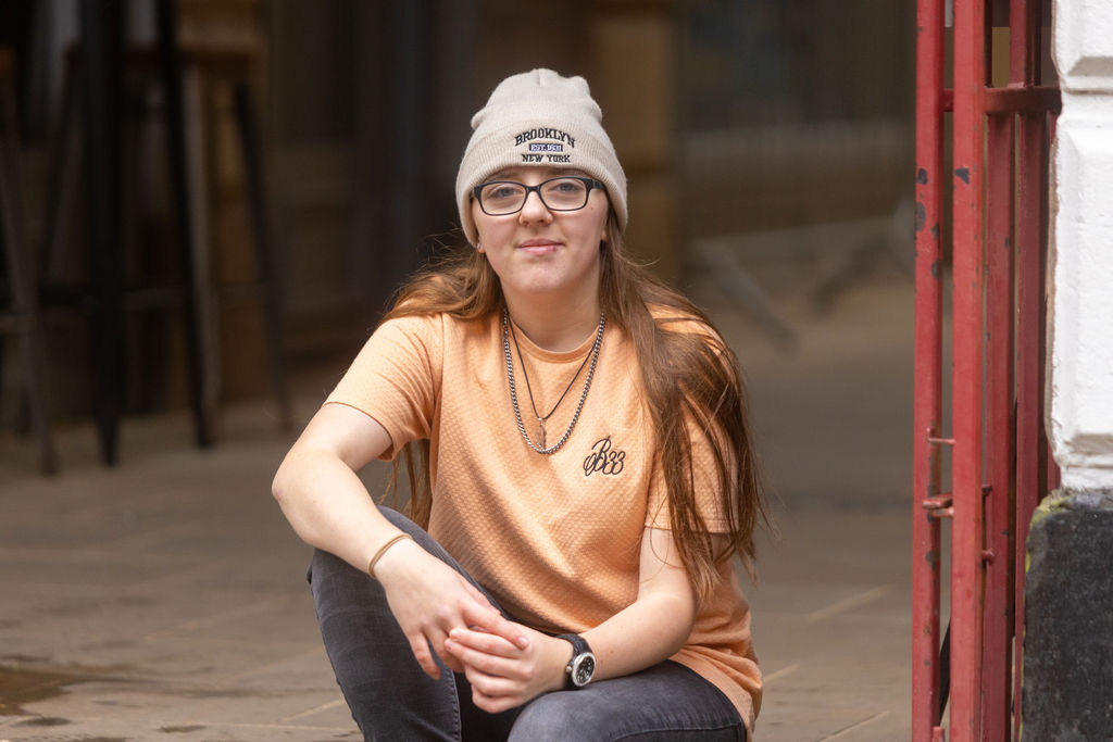 Young girl with glasses wearing a beanie hat and orange t-shirt sat with her arms rested on her legs.