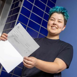 Student with a blue hair and blue t-shirt hiolding a document with exam results.