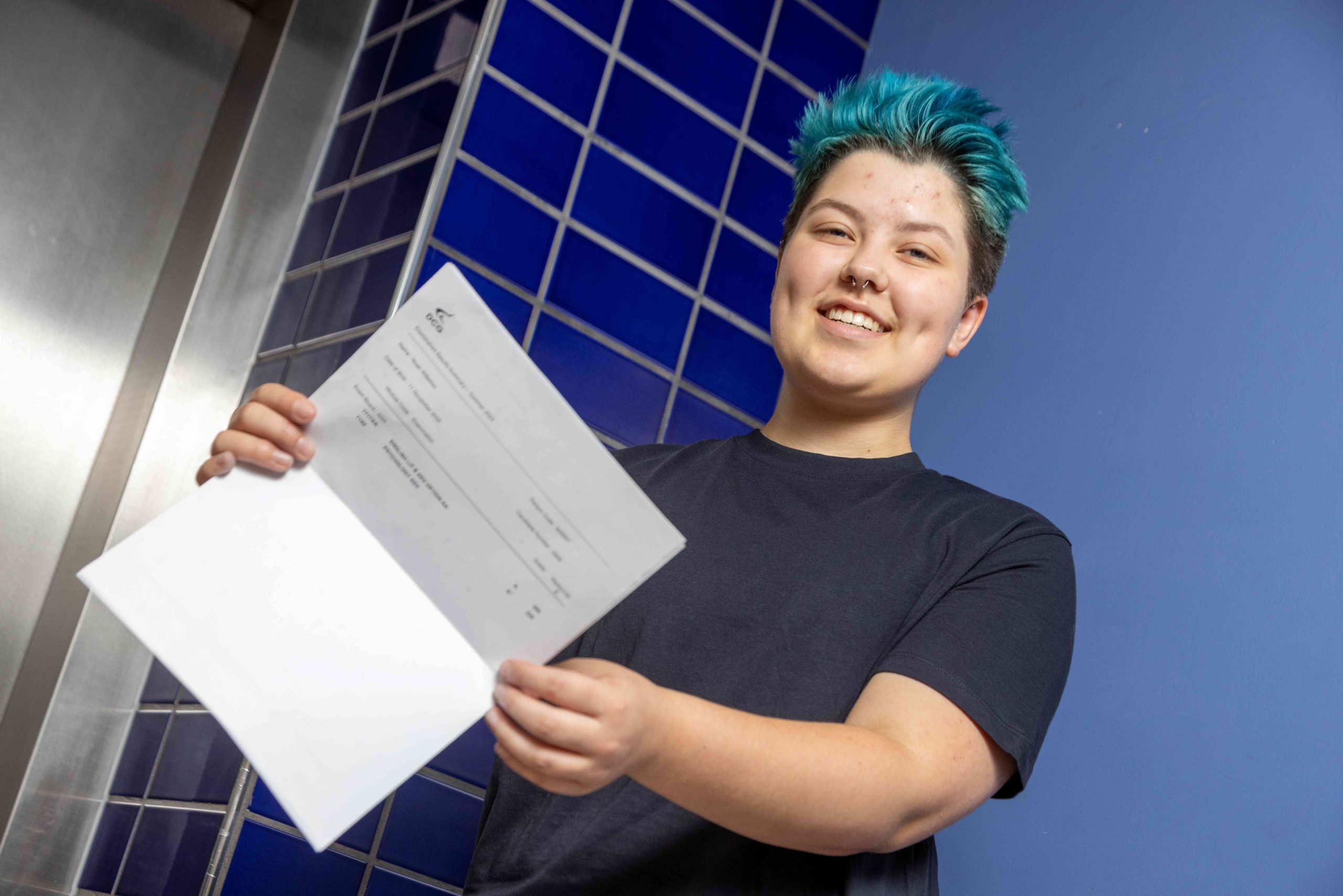 Student with a blue hair and blue t-shirt hiolding a document with exam results.