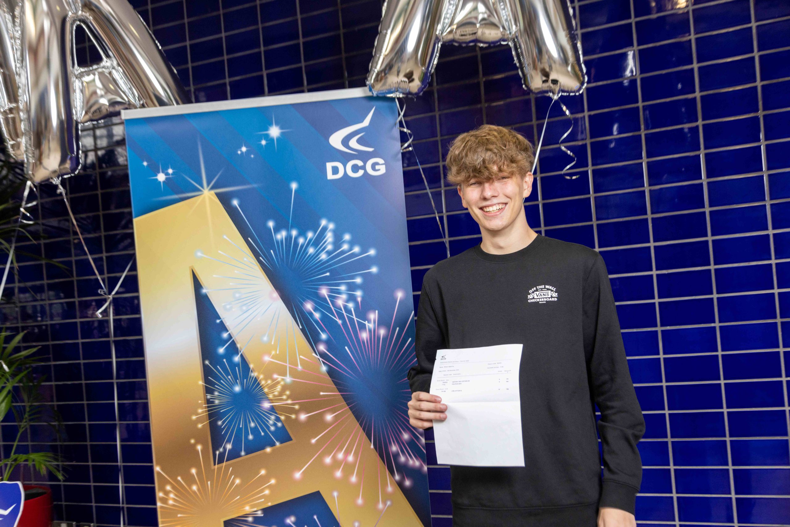 Young male with brown hair wearing a black sweater, holding a document with exam results on.