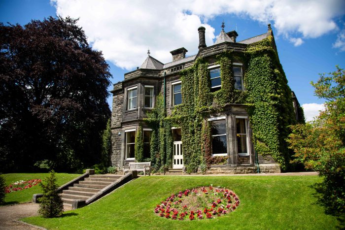 Large old building with covered in green leaves, surrounded by big trees with purple and green leaves. A bed of red flowers in the foreground.