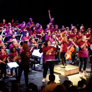 A large group of school & college students and adults performing with various instruments and singing.