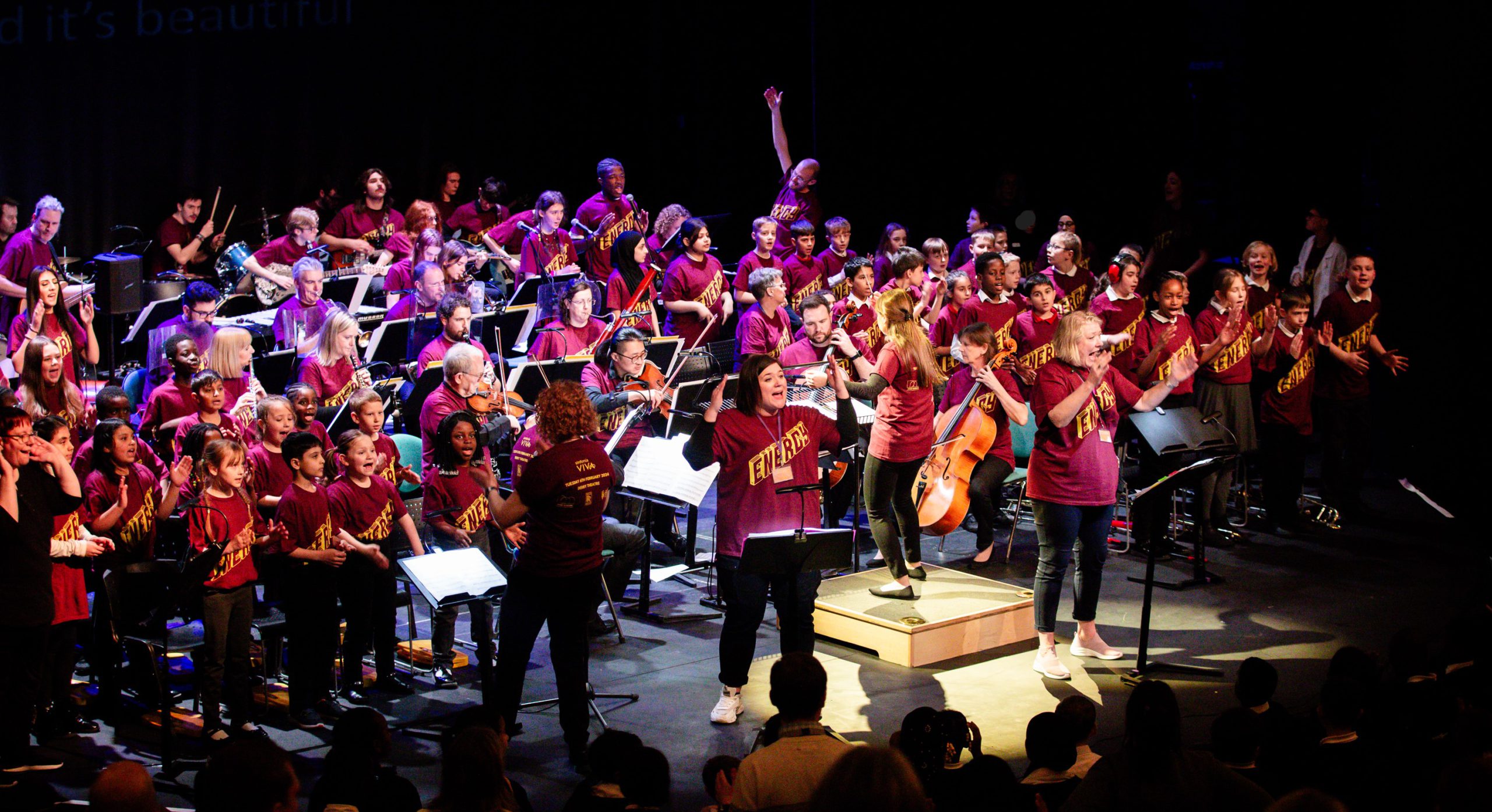 A large group of school & college students and adults performing with various instruments and singing.