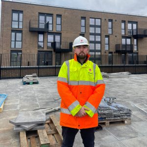 Higher Apprentice Tom wearing a yellow and orange hi-viz coat stood on a construction site.