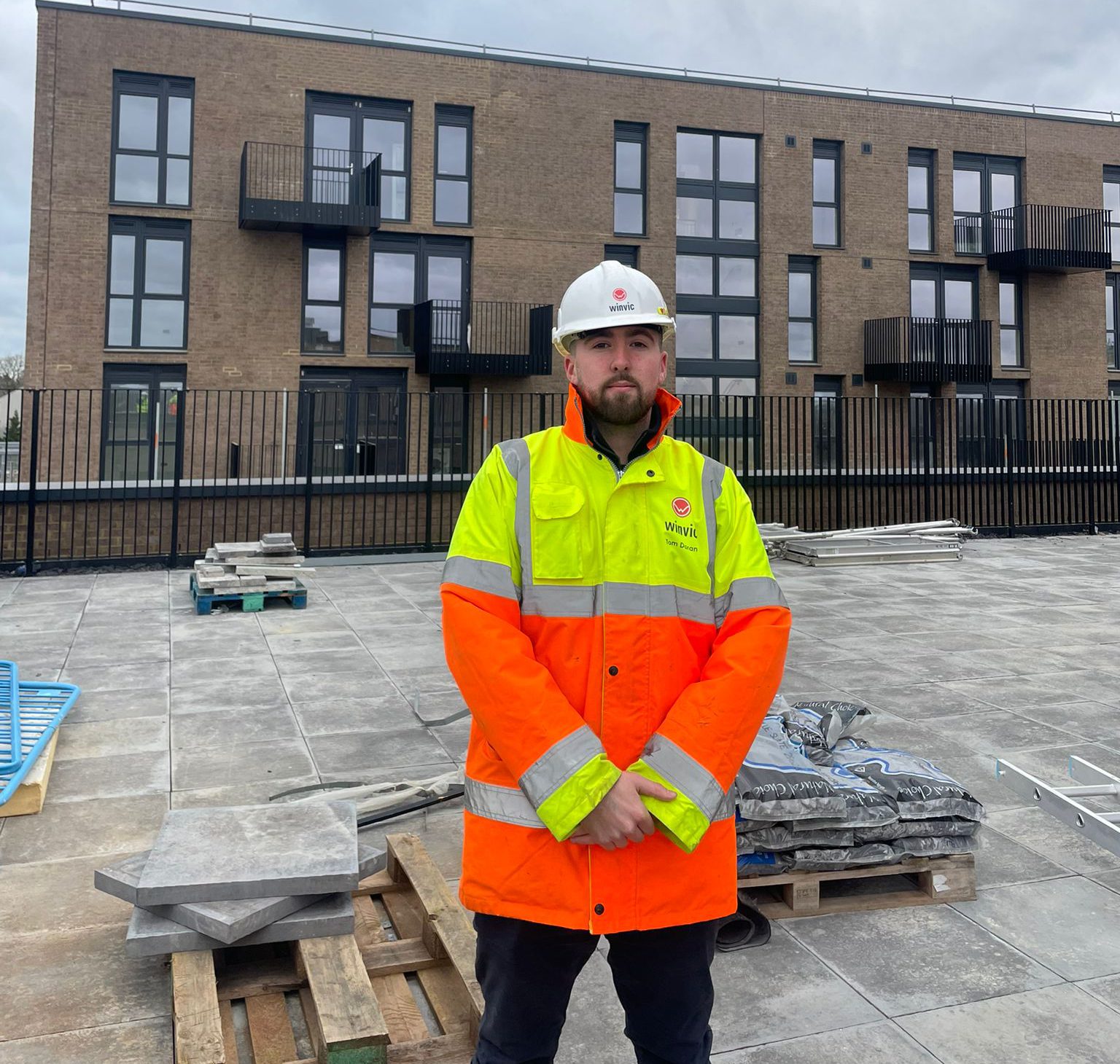 Higher Apprentice Tom wearing a yellow and orange hi-viz coat stood on a construction site.