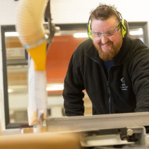 Teacher with a beard wearing goggles and ear protectors whilst operating machinery.