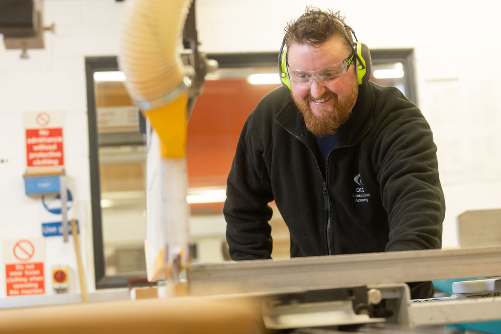 Teacher with a beard wearing goggles and ear protectors whilst operating machinery.