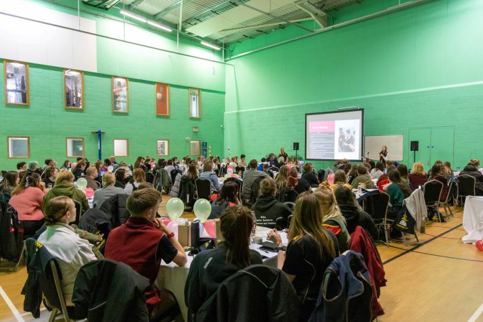 A group of students sat in a big sports hall with green walls.