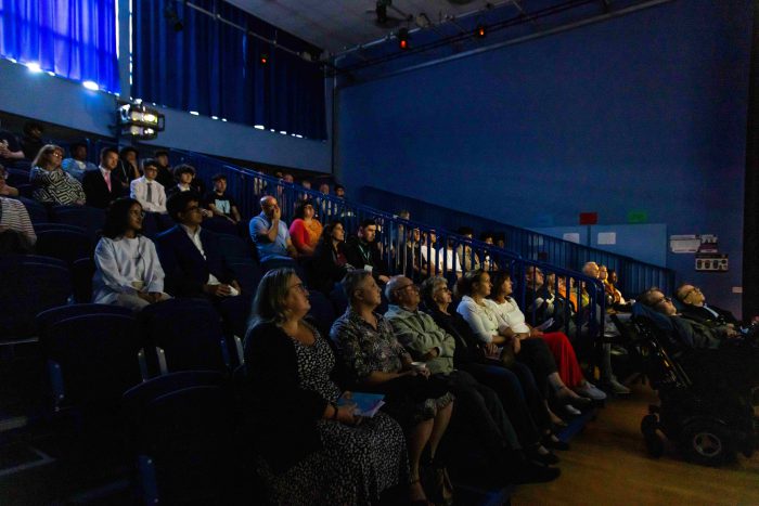 Spectators in a small theatre watching a large screen.
