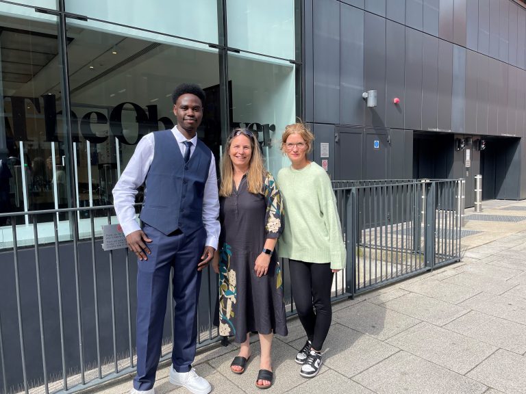 A male and female student stood either side of their teacher outside of a black building.