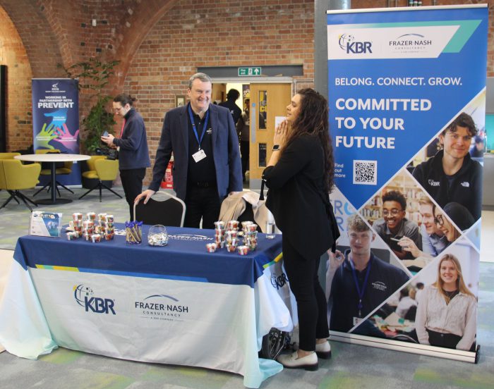 Two people stood behind by a stall talking to each other.