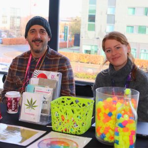 Two people wearing lanyards sat at a stall with a display.