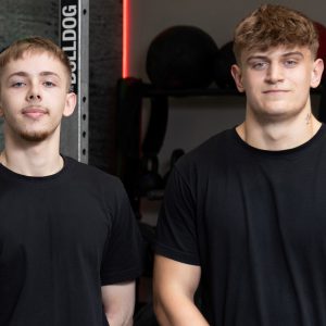 Two people wearing black t-shirts stood near weights in a gym.
