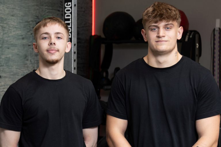 Two people wearing black t-shirts stood near weights in a gym.