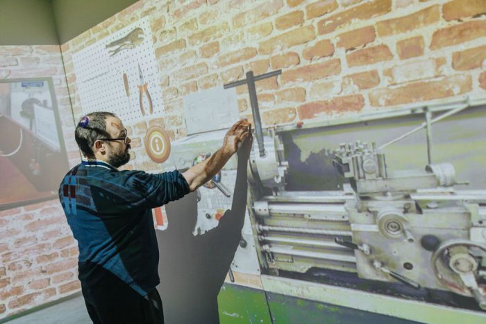 Person interacting with a large projected image of a workshop wall featuring tools and a metal lathe machine, demonstrating technical equipment in an industrial training environment.