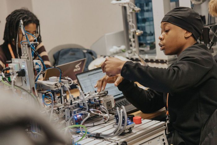 Person working on an advanced robotics or automation system with multiple components and wiring, seated at a desk with a laptop in a technical training environment.
