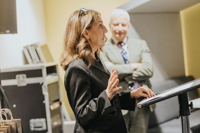 Speaker standing at a podium delivering a presentation during an event, with another person standing in the background near technical equipment.