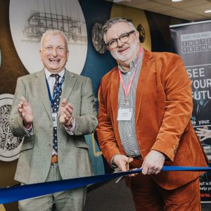 Two individuals holding a large blue ribbon during an official ribbon-cutting ceremony at East Midlands Institute of Technology, with a promotional banner in the background.