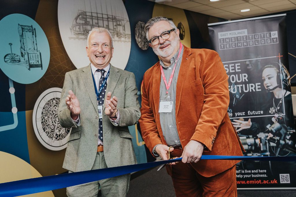 Two individuals holding a large blue ribbon during an official ribbon-cutting ceremony at East Midlands Institute of Technology, with a promotional banner in the background.
