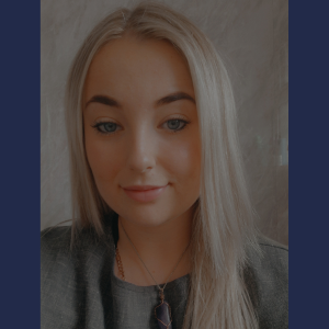 Person with long, straight, light blonde hair wearing a dark gray top and a necklace with a purple pendant, standing against a light textured background.