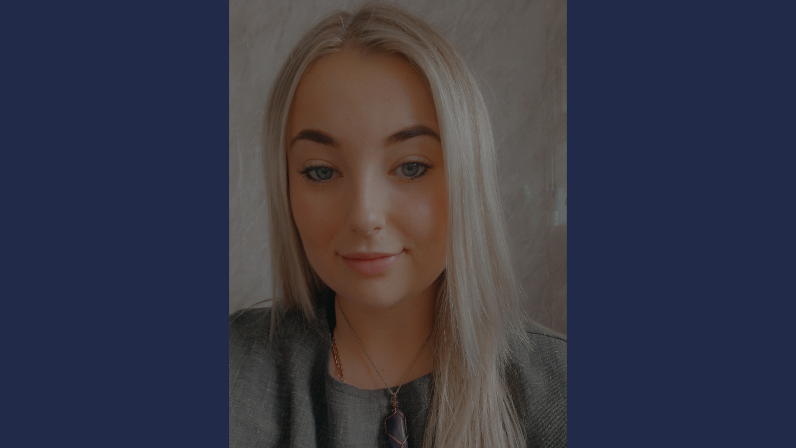 Person with long, straight, light blonde hair wearing a dark gray top and a necklace with a purple pendant, standing against a light textured background.