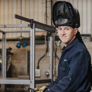 A person wearing a dark protective welding jacket and a raised welding helmet, seated in a workshop with metal frames, tools, and equipment in the background.