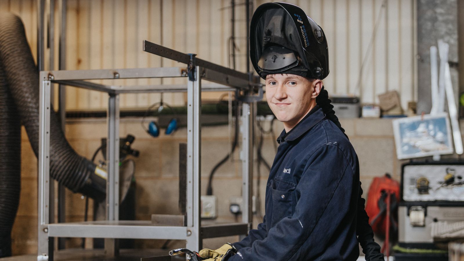 A person wearing a dark protective welding jacket and a raised welding helmet, seated in a workshop with metal frames, tools, and equipment in the background.