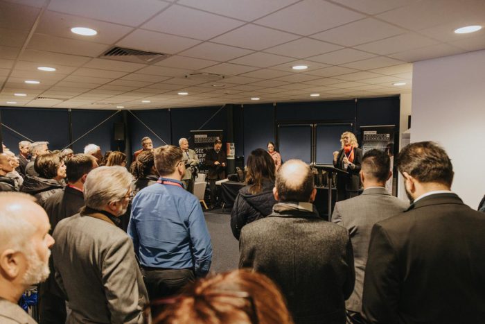 Audience gathered in a conference room listening to a speaker at a podium during an event for East Midlands Institute of Technology, with banners displayed around the room.