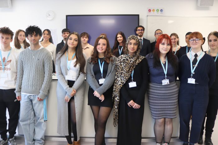 Group of T Level students standing together in a classroom, wearing light blue lanyards and ID badges, with a large screen and whiteboard in the background.