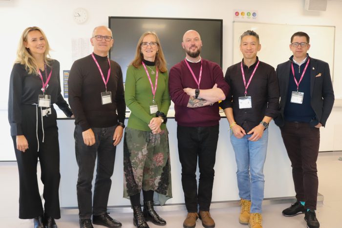 Group of T Level Dragons' Den event judges standing in a classroom, wearing pink lanyards and ID badges, positioned in front of a large screen and whiteboard.