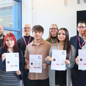 T Level ambassadors holding certificates in front of a display screen and signage, with others standing alongside them during a college T Level event.