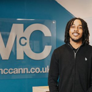 Person standing indoors in front of a blue wall with a large acrylic sign displaying the letters “Mc” and the website “fpmccann.co.uk.” The individual is wearing a black zip-up sweater with a small embroidered logo on the chest. A light-colored counter and a framed picture are visible in the background.