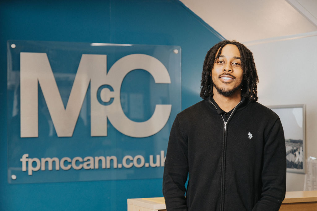 Person standing indoors in front of a blue wall with a large acrylic sign displaying the letters “Mc” and the website “fpmccann.co.uk.” The individual is wearing a black zip-up sweater with a small embroidered logo on the chest. A light-colored counter and a framed picture are visible in the background.