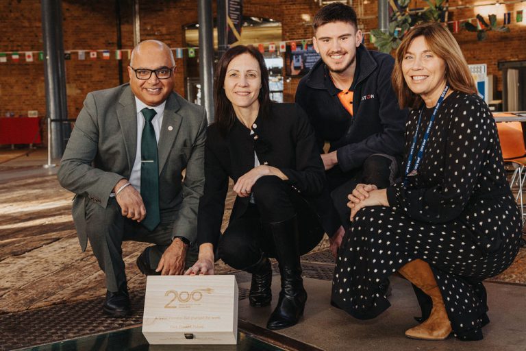 Four individuals are kneeling indoors around a wooden time capsule box engraved with the Railway 200 logo and the words “Rail Time Capsule.” The setting appears to be a spacious hall with brick walls, industrial-style pillars, and colorful flags hanging in the background. The group is dressed in formal and smart-casual attire, and the box is positioned prominently in the center of the image.