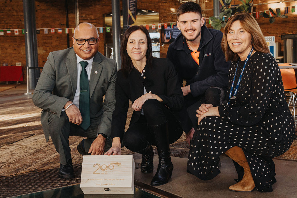 Four individuals are kneeling indoors around a wooden time capsule box engraved with the Railway 200 logo and the words “Rail Time Capsule.” The setting appears to be a spacious hall with brick walls, industrial-style pillars, and colorful flags hanging in the background. The group is dressed in formal and smart-casual attire, and the box is positioned prominently in the center of the image.