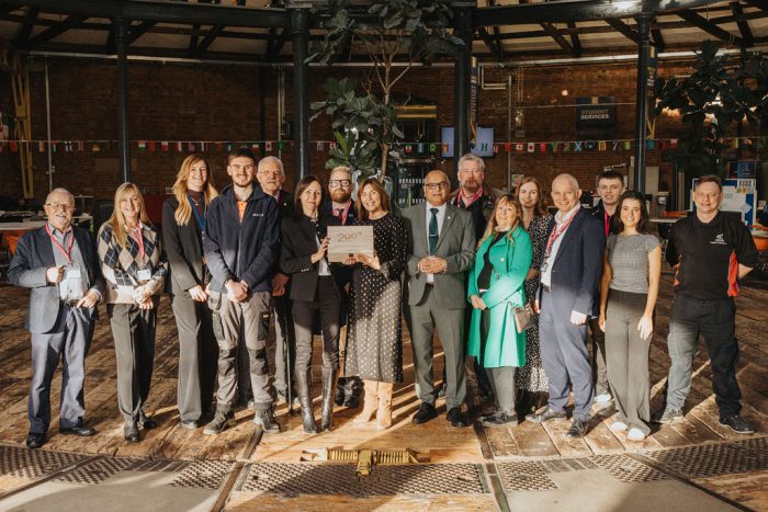 A group of people standing together indoors in a large hall with brick walls and industrial-style beams. They are positioned in a semi-circle on a wooden floor, holding a wooden time capsule box engraved with the Railway 200 logo. The background features colorful bunting, signage, and potted plants, creating a celebratory atmosphere.