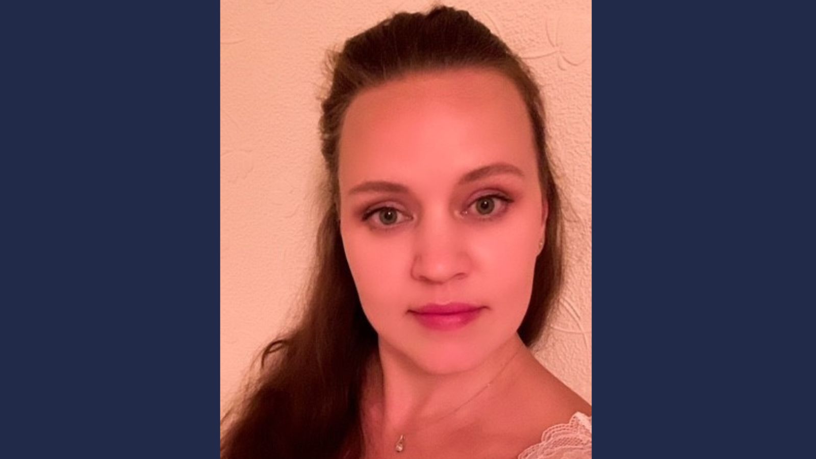 Person wearing a white top with lace detailing on one shoulder, standing against a light-coloured textured wall.