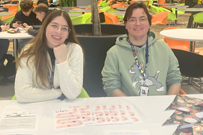 Two individuals seated at a table displaying printed sheets and cards labeled “12 DAYS” along with other informational materials. The setting is an open indoor area with multiple tables and bright green and orange chairs in the background, indicating a college or community event.