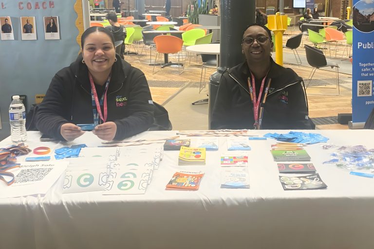 Two individuals sit behind a table covered with informational materials, wristbands, and leaflets in a bright indoor space with colorful chairs in the background. Items on the table include blue gloves, brochures, and a water bottle, suggesting a promotional or awareness stall.