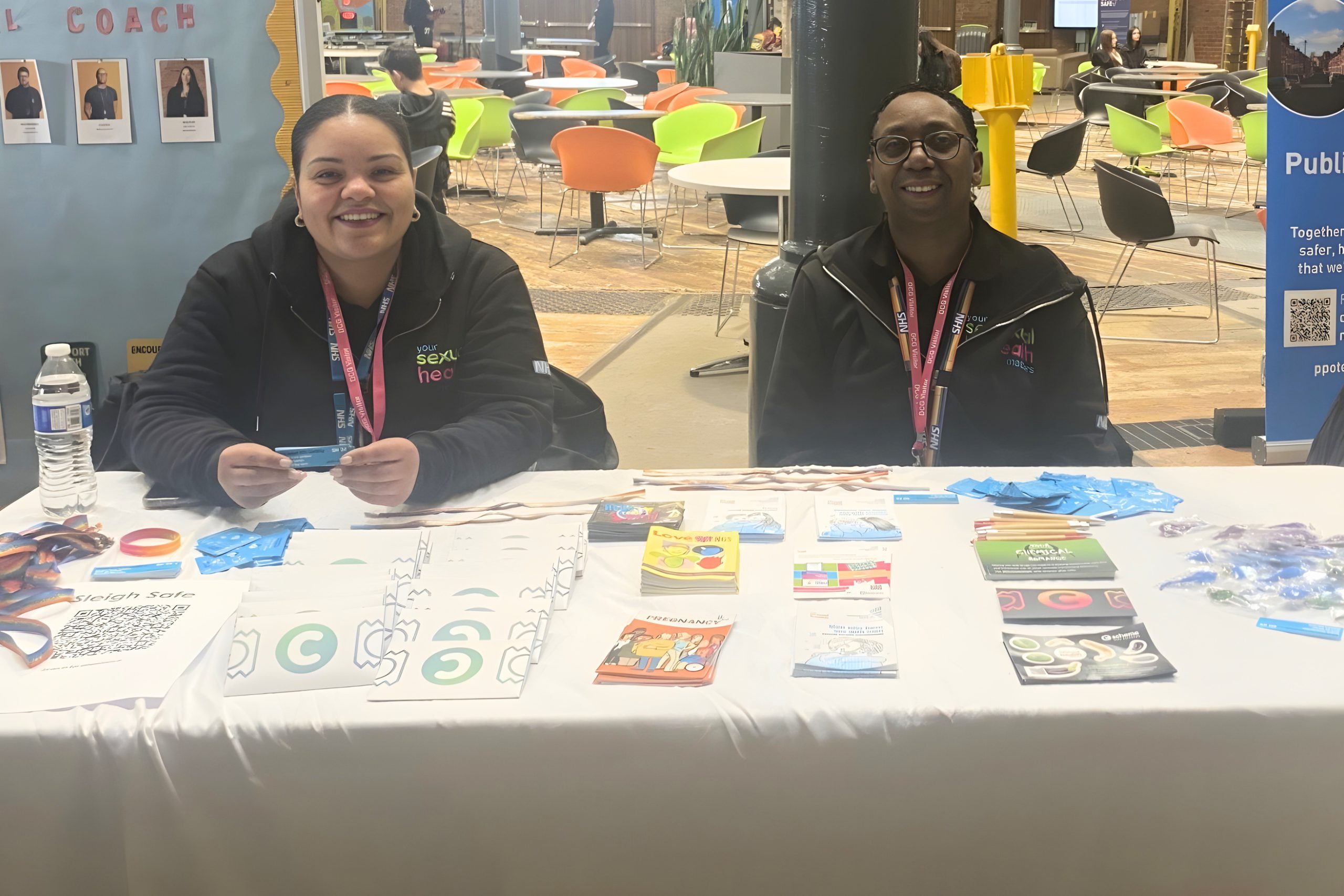 Two individuals sit behind a table covered with informational materials, wristbands, and leaflets in a bright indoor space with colorful chairs in the background. Items on the table include blue gloves, brochures, and a water bottle, suggesting a promotional or awareness stall.