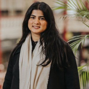 A person is standing indoors in front of a blurred background with architectural details and red pillars. The individual is wearing a black coat and a long cream-colored scarf, with dark hair falling over the shoulders. Green potted plants are visible to the right side of the image, adding a natural element to the scene.