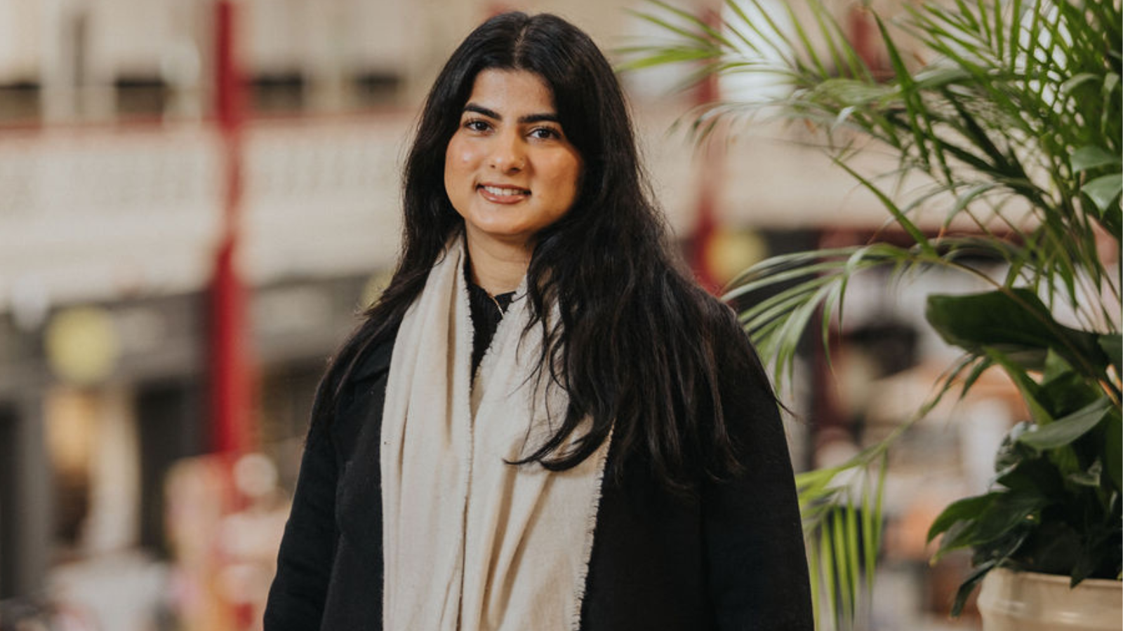 A person is standing indoors in front of a blurred background with architectural details and red pillars. The individual is wearing a black coat and a long cream-colored scarf, with dark hair falling over the shoulders. Green potted plants are visible to the right side of the image, adding a natural element to the scene.