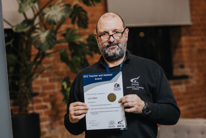 A person wearing a dark DCG-branded top holds a certificate titled “DCG Teacher and Assessor Award.” The certificate includes a gold seal and text indicating achievement in teaching and assessing. The background shows a brick wall and a large plant.